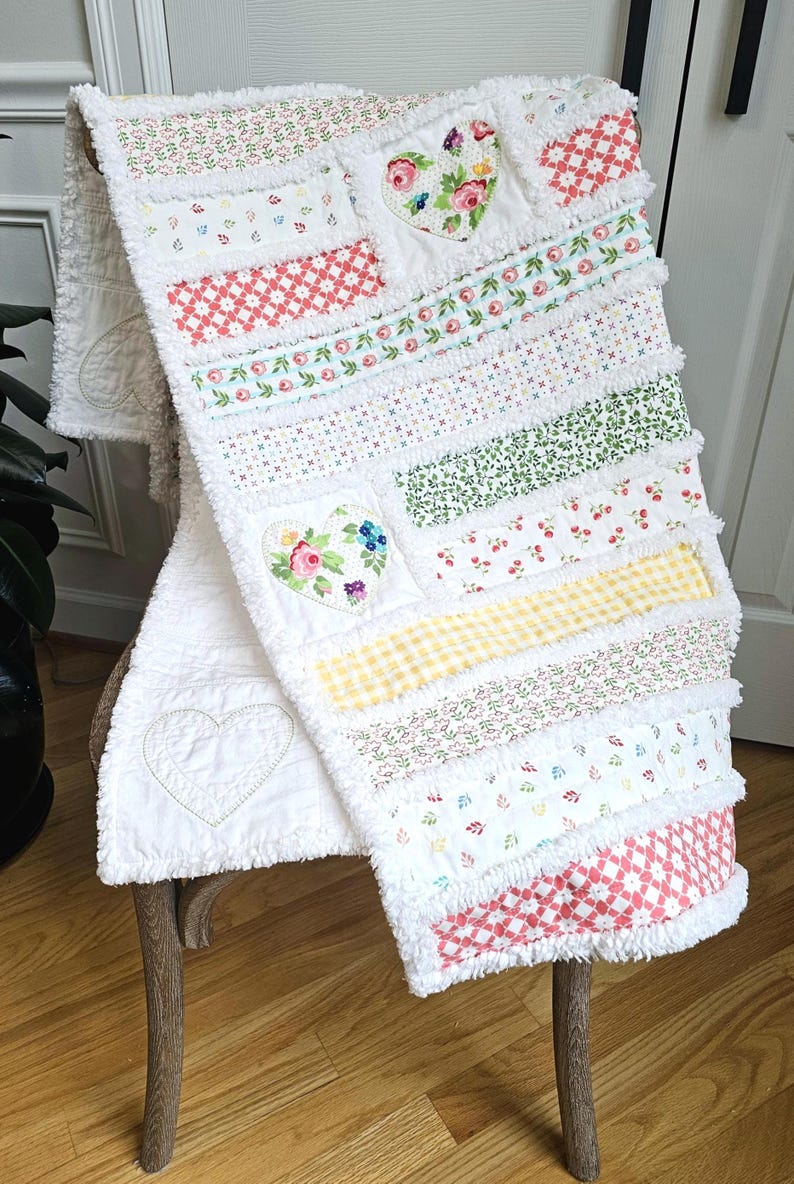 May include: A rag quilt featuring horizontal strips of white fabric with various floral, gingham, and geometric patterns in red, green, yellow, and blue. The quilt includes two fabric hearts with floral prints and one stitched heart outline.