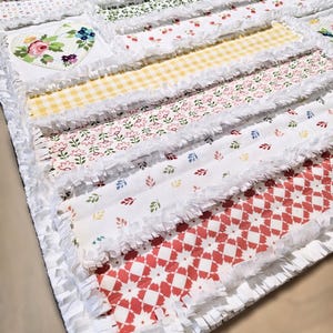 May include: A rag quilt featuring horizontal fabric strips with frayed white edges. The quilt displays various patterns including yellow gingham, red geometric shapes, floral prints, and a white square patch with a colorful floral heart design.