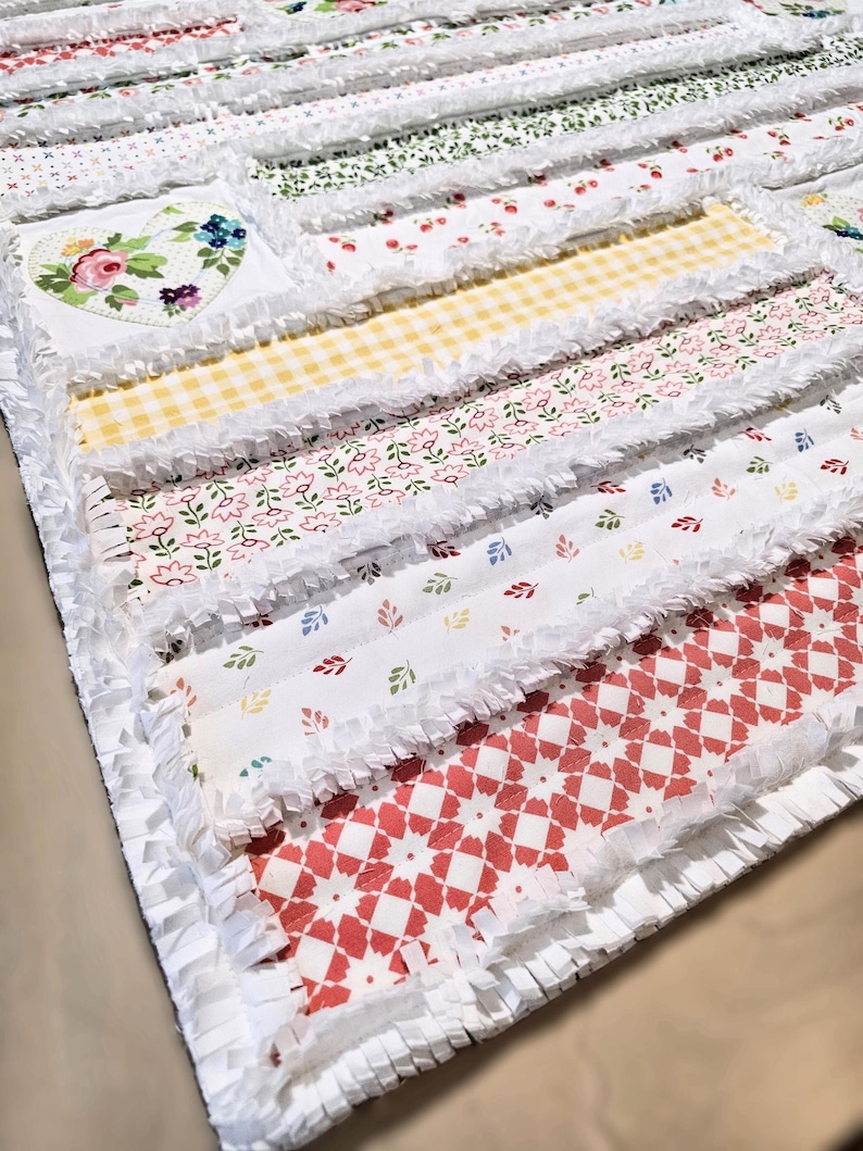 May include: A rag quilt featuring horizontal fabric strips with frayed white edges. The quilt displays various patterns including yellow gingham, red geometric shapes, floral prints, and a white square patch with a colorful floral heart design.