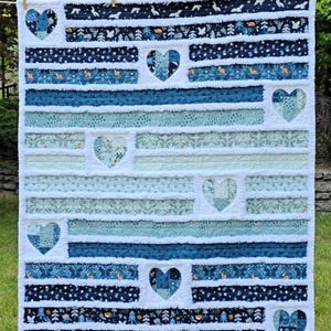 May include: A rectangular rag quilt featuring horizontal fabric strips in shades of navy blue, teal, and light blue. The quilt is decorated with seven heart-shaped patches integrated into the design, showcasing various woodland-themed patterns.