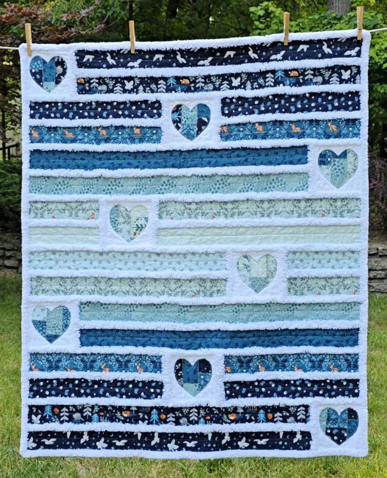 May include: A rectangular rag quilt featuring horizontal fabric strips in shades of navy blue, teal, and light blue. The quilt is decorated with seven heart-shaped patches integrated into the design, showcasing various woodland-themed patterns.
