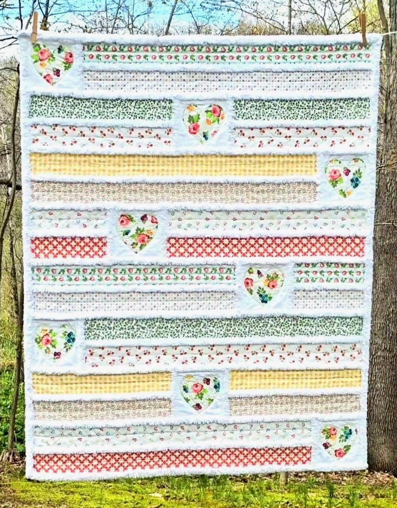 May include: A rectangular rag quilt featuring horizontal fabric strips in various floral, gingham, and geometric patterns. The quilt is accented with multiple heart-shaped fabric appliqu&eacute;s featuring floral designs, all separated by frayed white seams.