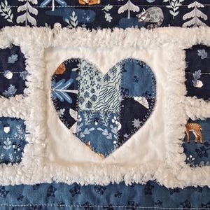 May include: A patchwork quilt featuring a central heart applique made of various blue and white patterned fabrics. The quilt is constructed with white fuzzy fabric borders between squares that display woodland motifs like deer, raccoons, and acorns.