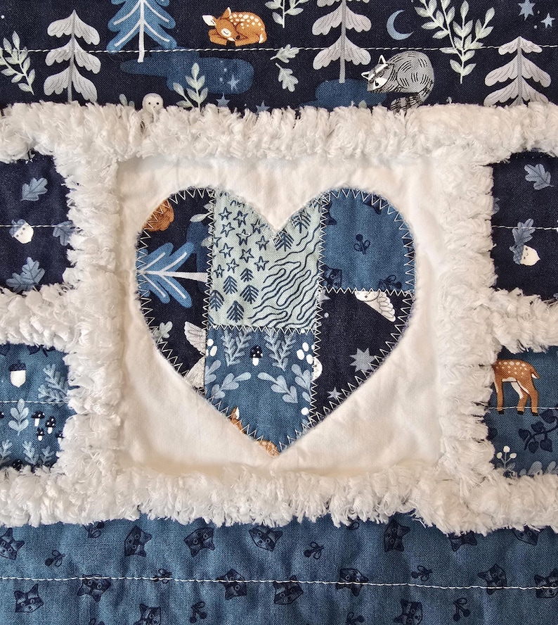 May include: A patchwork quilt featuring a central heart applique made of various blue and white patterned fabrics. The quilt is constructed with white fuzzy fabric borders between squares that display woodland motifs like deer, raccoons, and acorns.