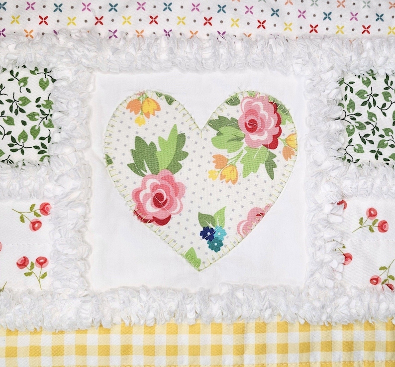 May include: A patchwork quilt featuring a central heart-shaped fabric appliqu&eacute; with pink roses and yellow flowers on a white dotted background. The quilt includes white frayed-edge seams, green leaf patterns, red berry prints, and a yellow gingham border.