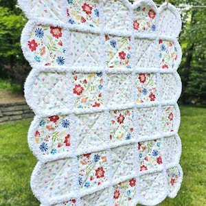 May include: A white floral quilt with red, blue, and yellow flowers is hanging on a clothesline. The quilt has a scalloped edge with a fluffy white trim. The quilt is made of small, square patches with a floral pattern.