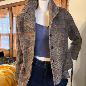May include: A plaid jacket in shades of gray, black, and red, displayed on a mannequin. The jacket has a wide collar, button closure, and a matching belt. The mannequin is wearing a blue top and dark blue jeans.