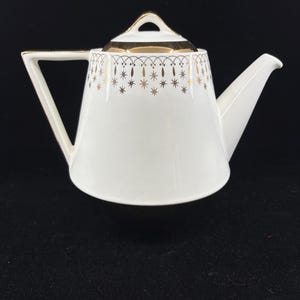 May include: White porcelain teapot with a gold geometric design. The teapot has a triangular handle and a spout. The lid is also white with gold trim. The teapot is on a black surface.