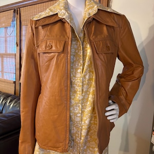 May include: A tan leather jacket with two front pockets and a zipper closure. The jacket is worn over a yellow floral print shirt. The jacket has a wide collar and is displayed on a mannequin.