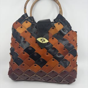 May include: A handcrafted leather tote bag with a unique woven design. The bag features a mix of black, brown, and tan leather pieces, secured with rivets. It has bamboo handles and a black leather flap with a gold-tone clasp.