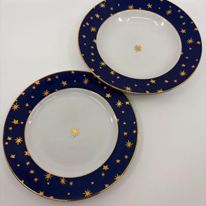 May include: Two white ceramic plates with a navy blue border adorned with gold stars. The plates have a gold star in the center and a gold trim. The plates are on a white background.