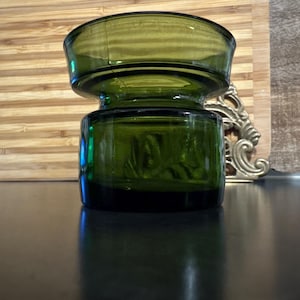 May include: A dark green glass candle holder with a wide, flared top and a cylindrical base. The glass has a slight texture and reflects light, creating highlights and shadows. The candle holder is sitting on a dark surface.