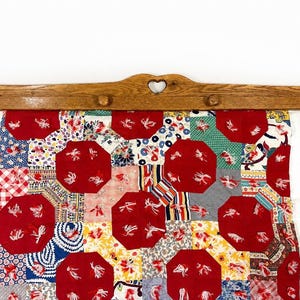 May include: A wooden quilt hanger featuring a central heart cutout and four pegs, suspending a patchwork quilt. The quilt displays a repeating pattern of red octagonal fabric patches interspersed with various colorful floral and geometric prints.