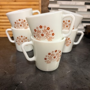 May include: A set of six white ceramic coffee cups with a brown floral design. Each cup has a handle and a flared shape. The cups are stacked and arranged on a dark surface, showcasing a vintage aesthetic.