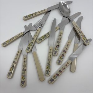 May include: A collection of vintage children's flatware with cream-colored handles decorated with colorful house and animal motifs. The set includes knives, forks, and a spoon, all with stainless steel blades and tines.