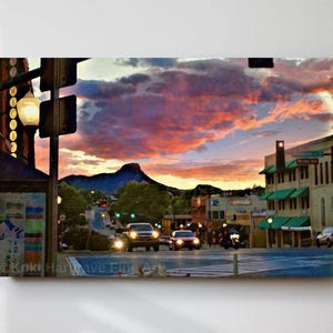 May include: A canvas print of a town scene at sunset. The sky is filled with vibrant pink and orange clouds. The street has cars and buildings with green awnings. A mountain is in the background.