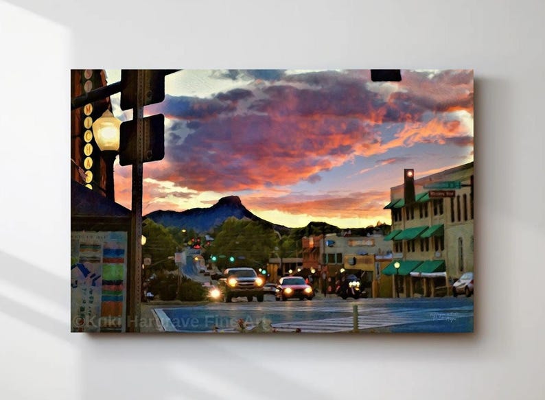 May include: A canvas print of a town scene at sunset. The sky is filled with vibrant pink and orange clouds. The street has cars and buildings with green awnings. A mountain is in the background.
