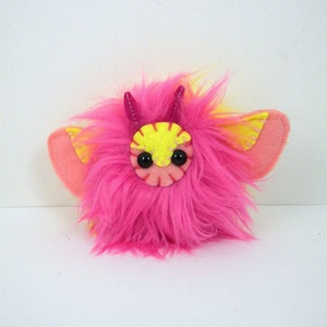 May include: A pink and yellow fuzzy plush toy with horns and wings. The toy has black eyes and a pink mouth.