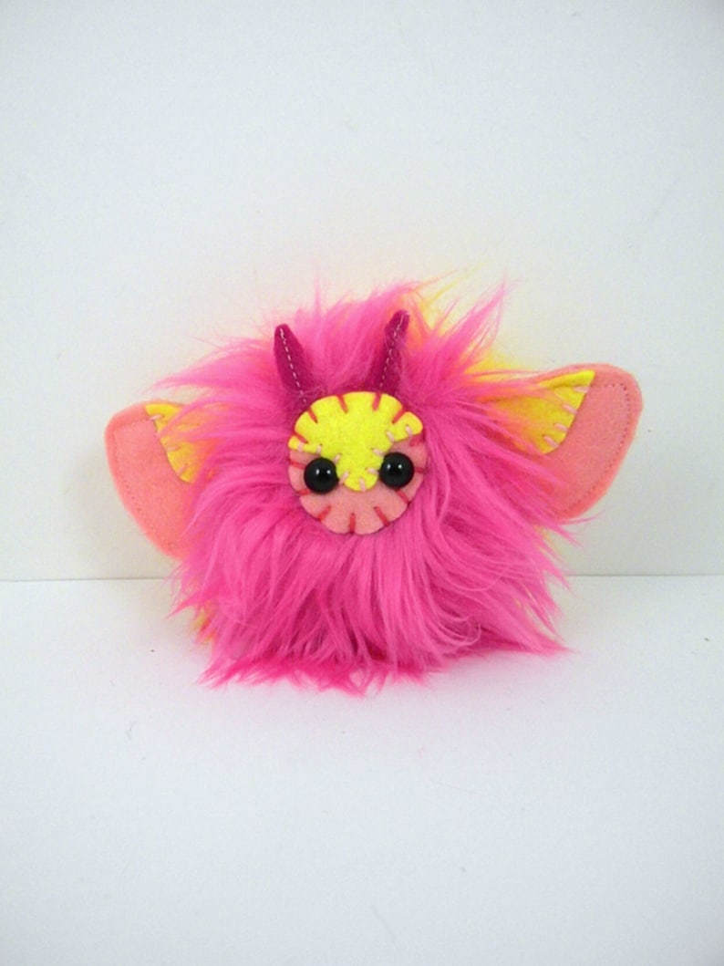 May include: A pink and yellow fuzzy plush toy with horns and wings. The toy has black eyes and a pink mouth.