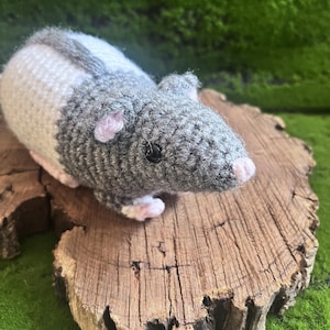 May include: A crocheted rat toy featuring a two-tone design with a dark gray head and front body section, transitioning into a white back section. The rat has small pink ears, a pink nose, black bead eyes, and light pink feet.