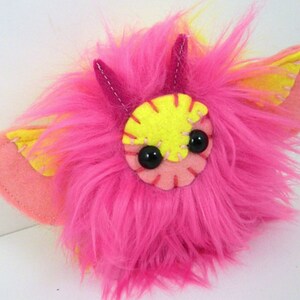 May include: A pink and yellow fuzzy plush toy with large black eyes and a stitched smile. The toy has small pink horns and yellow wings.