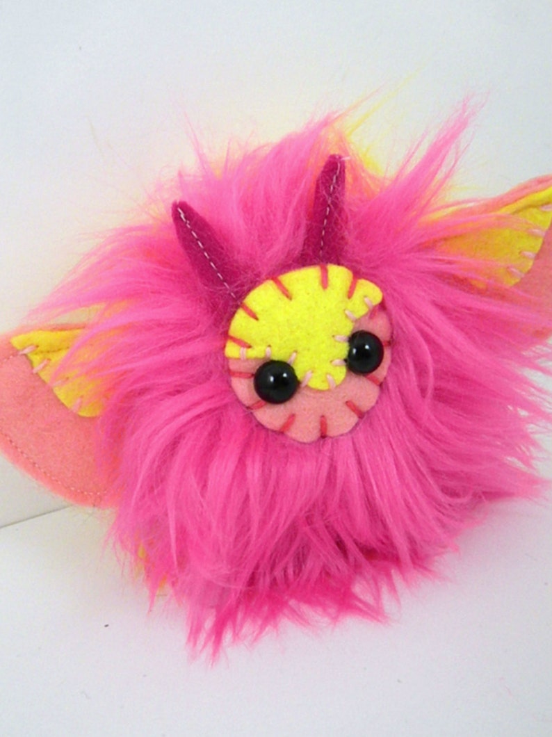 May include: A pink and yellow fuzzy plush toy with large black eyes and a stitched smile. The toy has small pink horns and yellow wings.