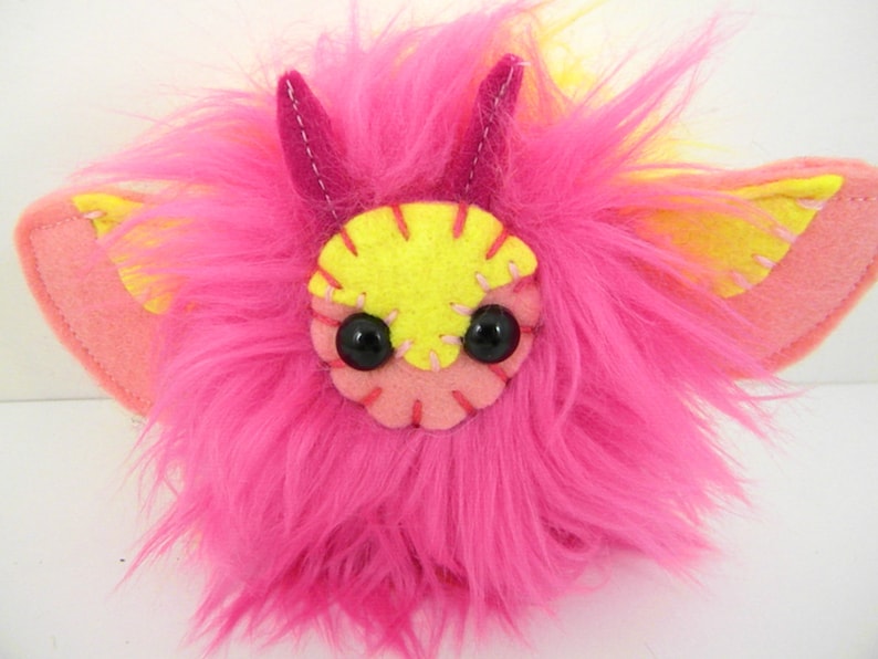 Pink and yellow faux fur plush moth toy with long pink fur, small black eyes, and pink and yellow stitched details. The moth has small pink antennae and large pink felt bat-like wings with yellow accents.  The handmade plush toy is a whimsical, cute creature, perfect for children or collectors of unique handmade toys.