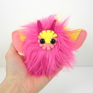 May include: A pink and yellow fuzzy plush toy with horns and wings. The toy has black button eyes and a stitched mouth.