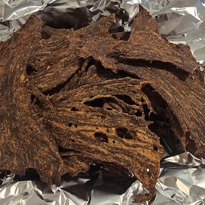 May include: A pile of dark brown, dehydrated beef jerky strips with a textured, fibrous surface and irregular edges. The dried meat pieces are stacked together, displaying a deep mahogany color with visible seasoning particles throughout the surface.