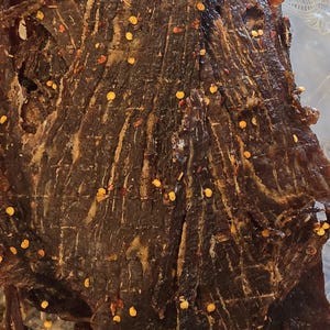 May include: A stack of dark brown, dried beef jerky strips seasoned with scattered red chili pepper flakes. The meat has a fibrous, textured appearance with visible grain lines and is contained within a clear plastic food storage container.