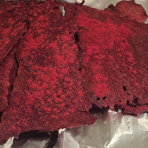 May include: Several thin, irregular pieces of dehydrated beet chips with a deep crimson and bright red color. The vegetable slices have a textured, brittle appearance with visible surface variations and small holes throughout the dried material.