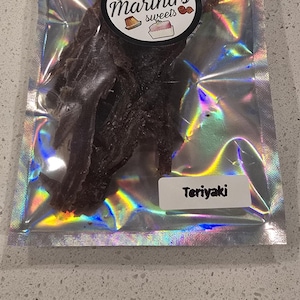May include: A holographic, resealable pouch containing dark brown, dried meat strips. A circular sticker at the top displays the text Doña Martha's sweets with illustrations of desserts. A white rectangular label at the bottom reads Teriyaki.