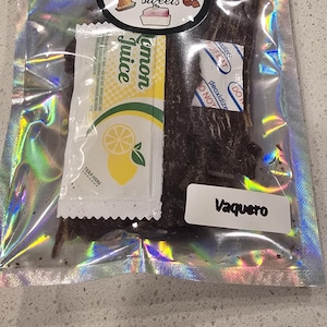 May include: A holographic pouch containing dark brown beef jerky, a small packet of lemon juice, and a white deoxidizer packet. The pouch features a circular logo for Doña Martha's sweets and a white label with the text Vaquero printed in black.