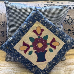 May include: A square cushion with a floral design. The cushion features a central square with a vase of flowers in red, yellow, and green. The border is a blue floral pattern. A second cushion with a blue and white checkered pattern is behind it.