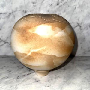 May include: A spherical ceramic vase with a textured, cracked rim and a rounded base. The vase has a blend of tan, brown, and white hues, creating a marbled effect. The design is organic and earthy.