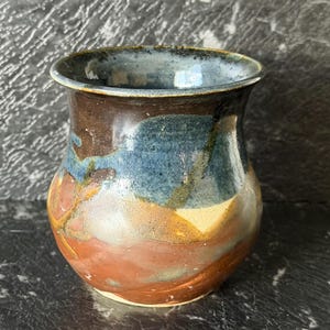 May include: A small, rounded ceramic vase or cup featuring a flared rim and a smooth, glazed finish. The exterior displays a blend of earthy colours, including deep brown, terracotta orange, and mottled blue tones, creating a variegated appearance.