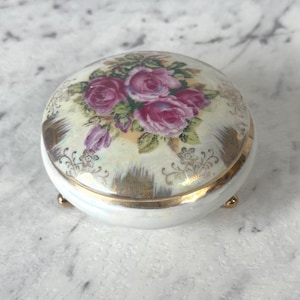 May include: A small, round, white porcelain trinket box with a lid featuring a floral design of pink roses and green leaves. The box has gold trim and rests on three small gold feet.