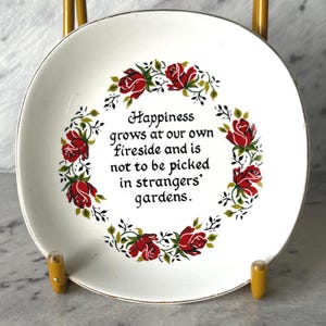 Op de afbeelding: Een rond wit decoratief keramieken bord met een centrale zwarte tekstinscriptie die luidt: Happiness grows at our own fireside and is not to be picked in strangers' gardens. De tekst is omringd door een cirkelvormige krans van rode rozen.