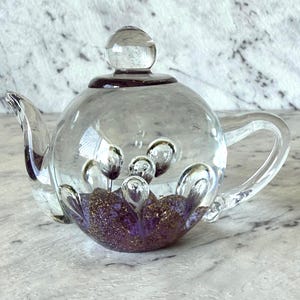 May include: A clear glass teapot with a rounded body, spout, and handle. The base features a purple and gold glitter design. The lid has a small, round knob. Inside the teapot are several clear, bubble-like shapes.