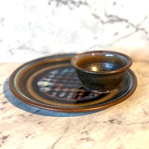 以下が含まれることがあります： 手作りの小さなセラミックボウルとソーサーのセット。ボウルはマッチするソーサーの上にあります。セットは、茶色と青色の配色とテクスチャパターンが特徴です。セットは、小さな部分を提供するのに使用される可能性があります。