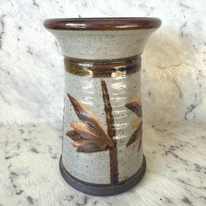 May include: A speckled ceramic vase with a wide rim and a dark brown base. The vase features a brown and tan leaf design and a brown band around the middle. The vase is approximately 18 cm tall.