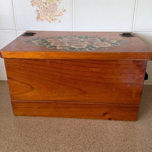 May include: A rectangular wooden storage chest with a polished medium-brown finish. The lid features a painted floral design with muted pink roses and green leaves. Two dark metal hinges are visible on the top edge of the wooden box.