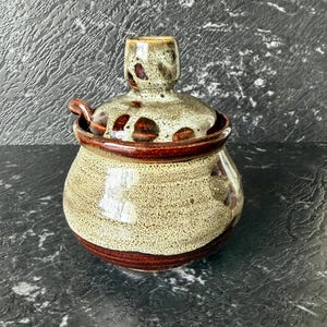 May include: A handmade ceramic jar with a lid. The jar is a rounded shape with a speckled beige and brown glaze. The lid has a small handle and is decorated with brown spots. The jar has a dark brown band around the base and rim.