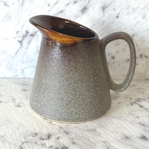 Può includere: Una brocca in ceramica con un corpo grigio strutturato e un bordo marrone. L'impugnatura è un anello curvo e il beccuccio è angolato per facilitare il versamento. La brocca ha un aspetto rustico e fatto a mano.