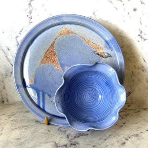 May include: A round ceramic plate in periwinkle blue with a speckled finish and a triangular copper-coloured metallic accent, paired with a smaller, matching periwinkle blue ceramic bowl featuring a scalloped edge and concentric circular ridges.
