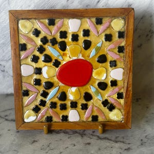 May include: A square decorative tile with a mosaic design. The tile has a wooden frame and a central red oval surrounded by yellow, black, pink, white, and blue shapes. It is supported by a small wooden stand.