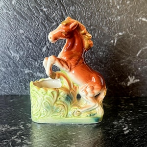 May include: A ceramic horse figurine in shades of brown, orange, and cream, rearing up from a green and yellow planter base. The planter has a detailed, leafy design. The horse has a detailed mane and tail.