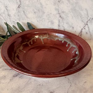 May include: A round, burgundy ceramic bowl with a wide rim. The interior has a decorative, dripped glaze in gold and white. The bowl is set against a marble-patterned background, suggesting a rustic or artisanal aesthetic. The bowl's diameter is approximately 25 cm.