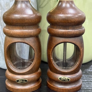 May include: Two wooden salt and pepper grinders with a clear glass window. The grinders are brown with a brass top and a base that reads "MADE IN JAPAN". The grinders have a classic, vintage design.