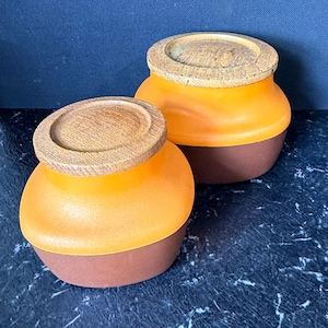 May include: Two small, round containers with lids. The containers have a brown base, an orange midsection, and a wooden lid. The containers are on a dark blue surface. The lids are round and made of light-coloured wood.
